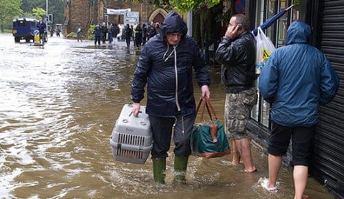 Äldre man i regnkläder och stövlar går med packning på översvämmad gata.