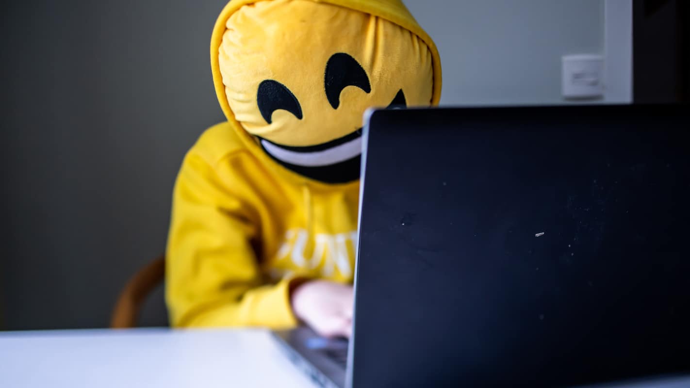 Person med emojikudde framför ansiktet sitter vid dator.