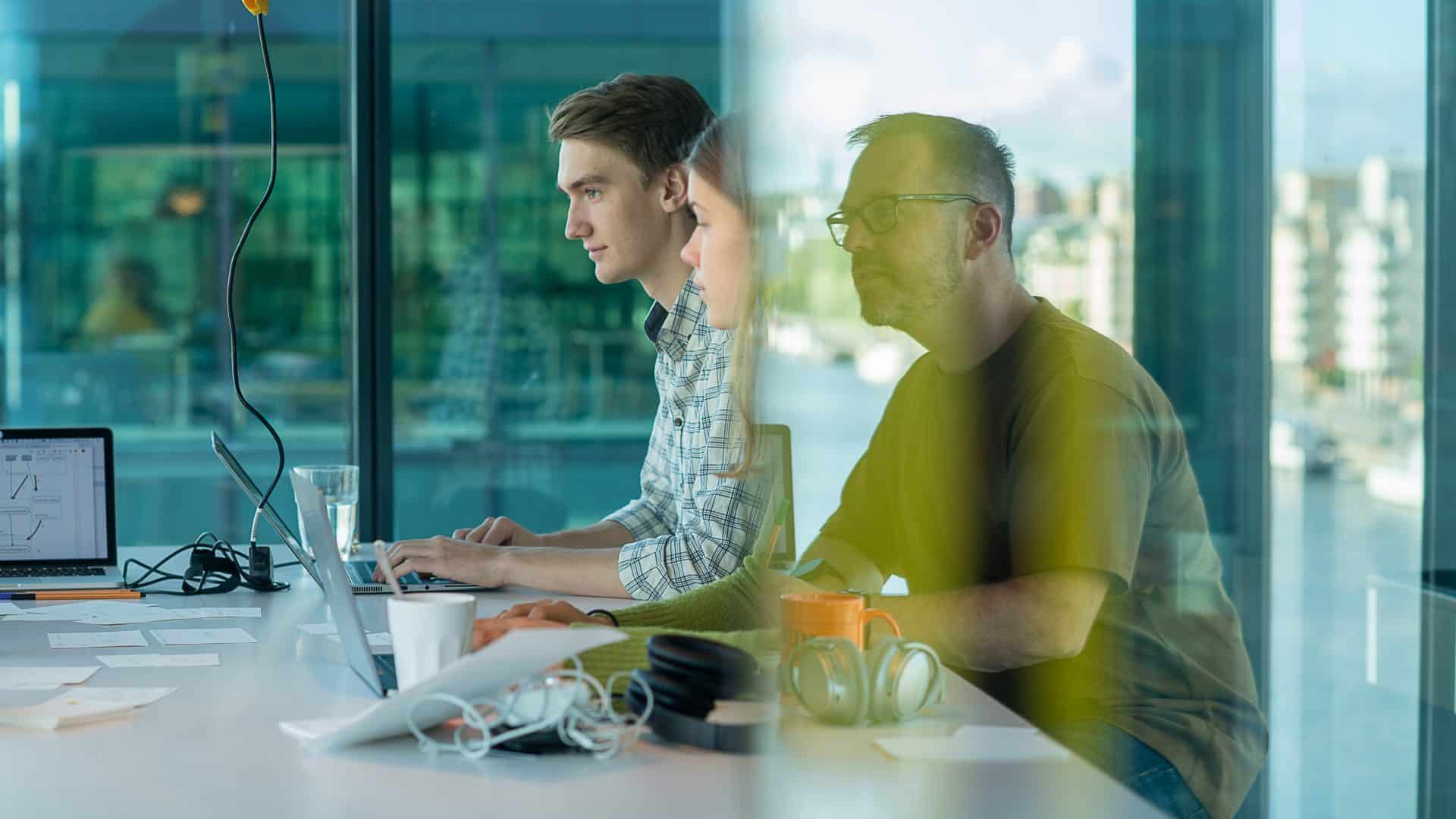 Tre kollegor sitter och arbetar med sina datorer vid ett bord på ett kontor med stora glasfönster.