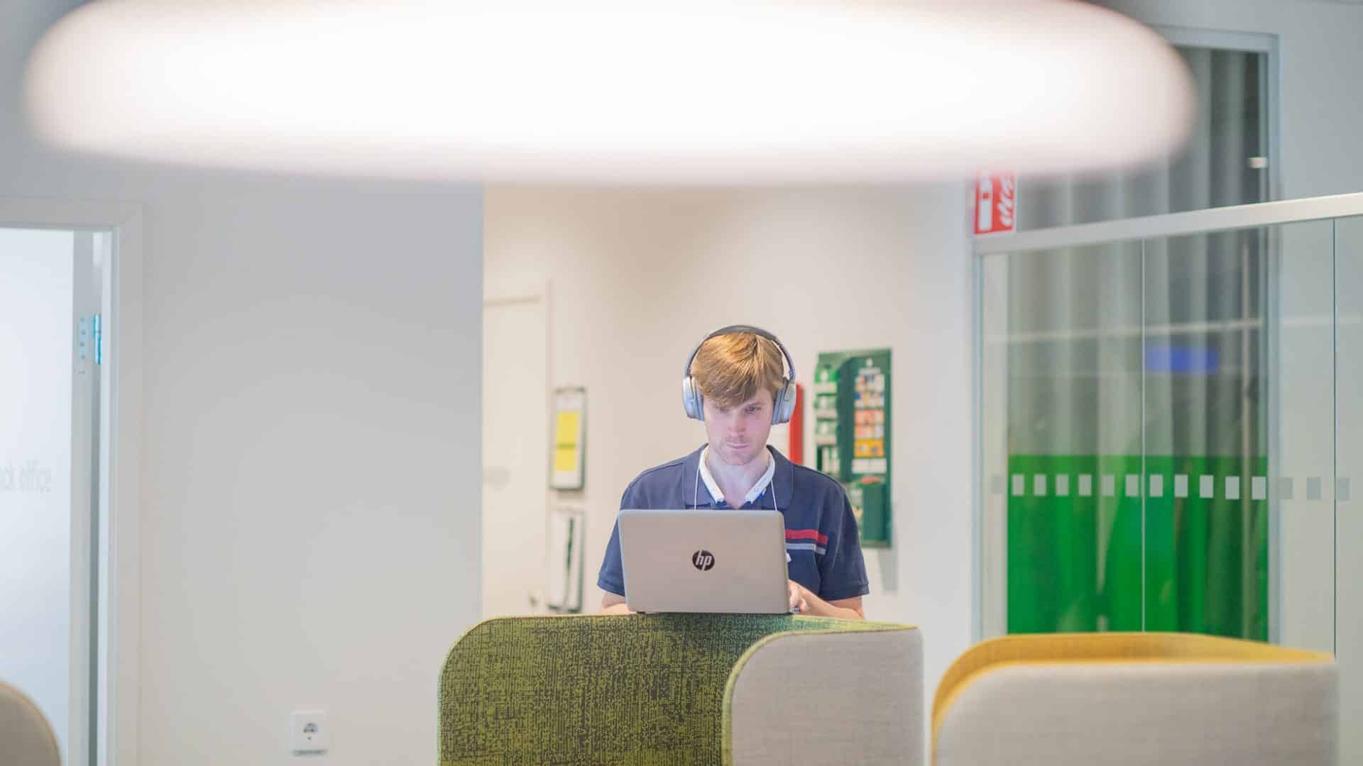 En man med hörlurar står upp och arbetar vid sin laptop.