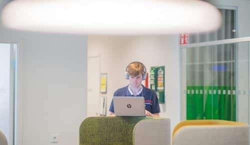 En man med hörlurar står upp och arbetar vid sin laptop.