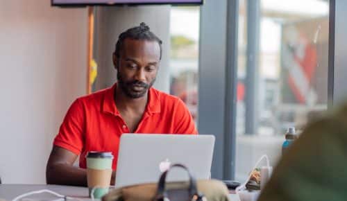 En man sitter i ett coworkingspace med en bärbar dator och en takeawaymugg kaffe