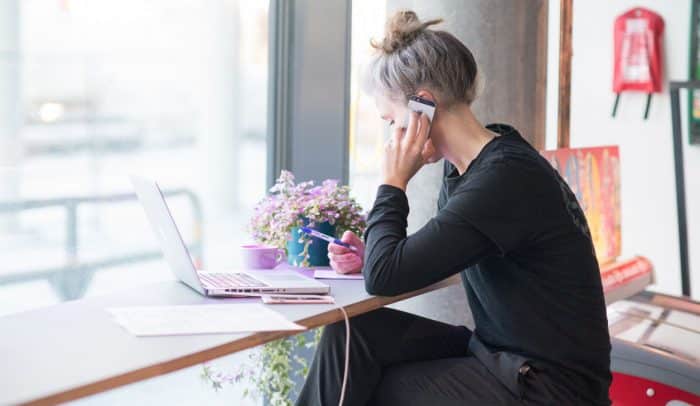 En kvinna sitter på ett kafé och pratar i telefon med en dator framför sig.