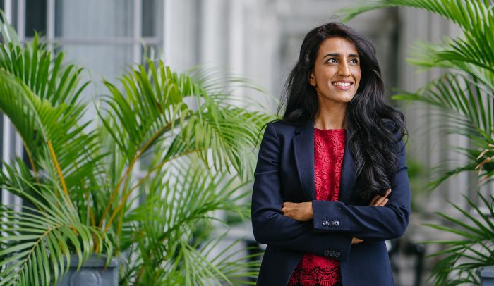Doktor Ayesha Khanna står på en innergård med växter i bakgrunden. Hon har kavaj på sig och tittar uppåt åt höger