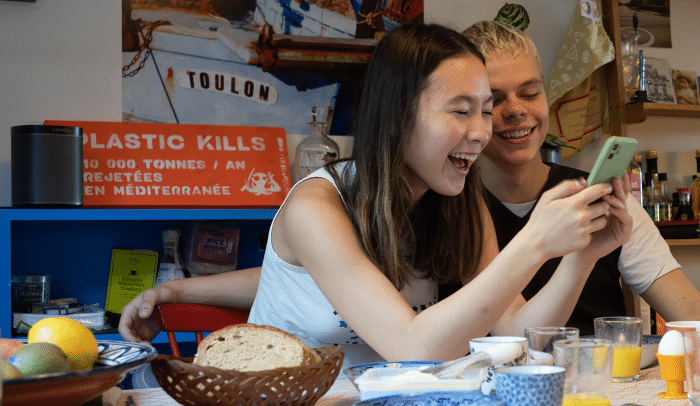 Två tonåringar sitter vid köksbordet och skrattar åt något de ser i mobilen.