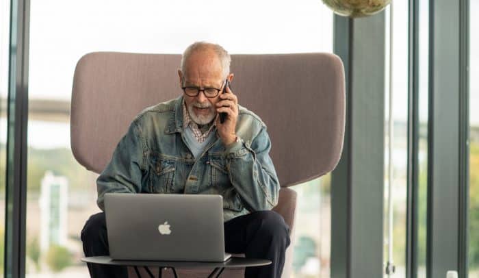 En äldre man med jeansjacka sitter vid sin dator och pratar i telefon.
