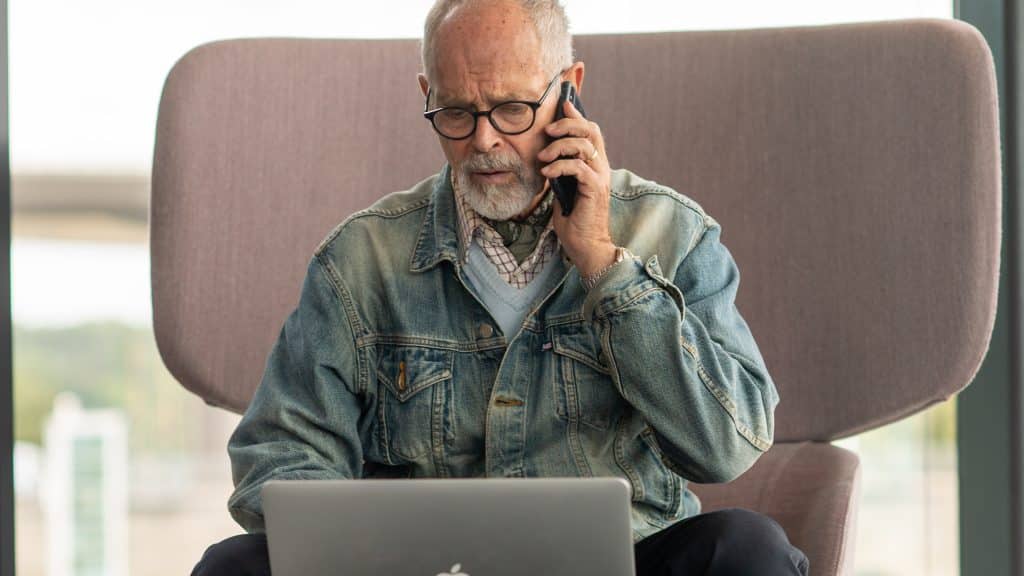 Äldre man sitter framför datorn och ringer ett telefonsamtal, orolig. Digitala samhällstjänster.