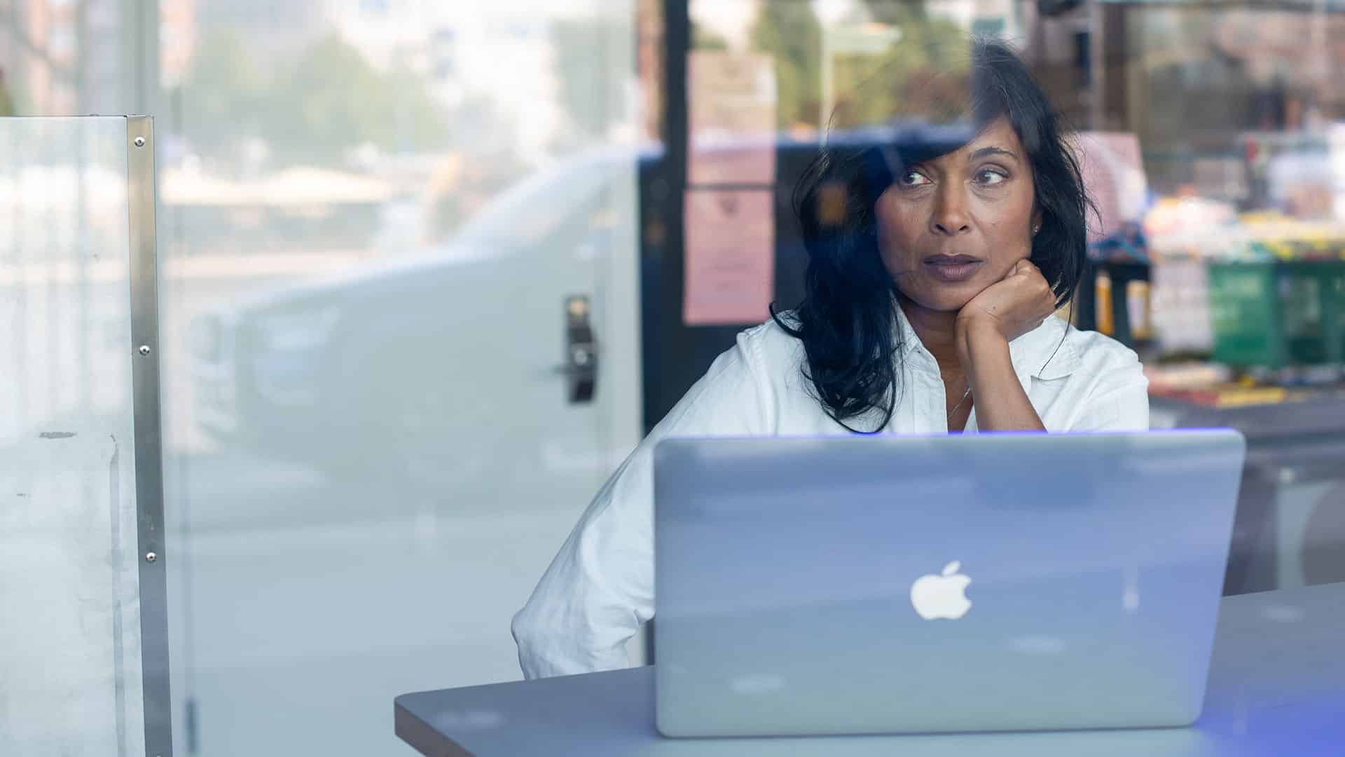 Kvinna sitter framför dator, tittar ut genom fönster.