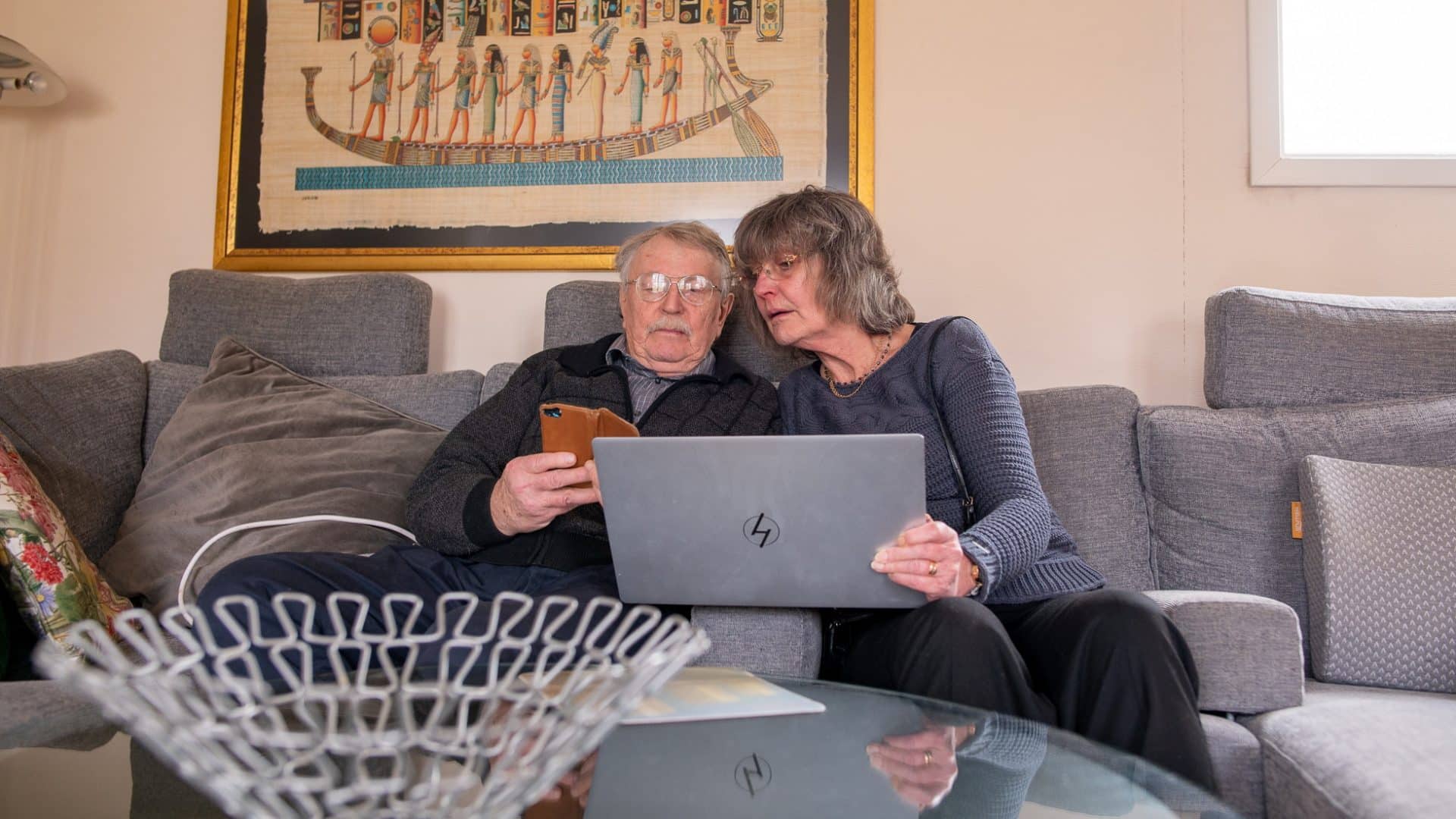 Man och kvinna sitter med mobil och bärbar dator i soffan.