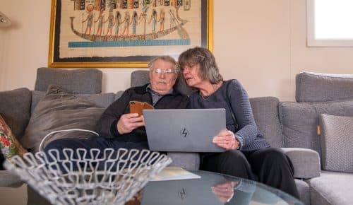 Man och kvinna sitter med mobil och bärbar dator i soffan.