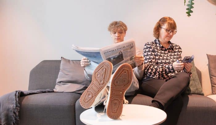 En ung kille och en äldre kvinna sitter i en soffa, kvinnan tittar på sin mobil, killen läser en tidning.