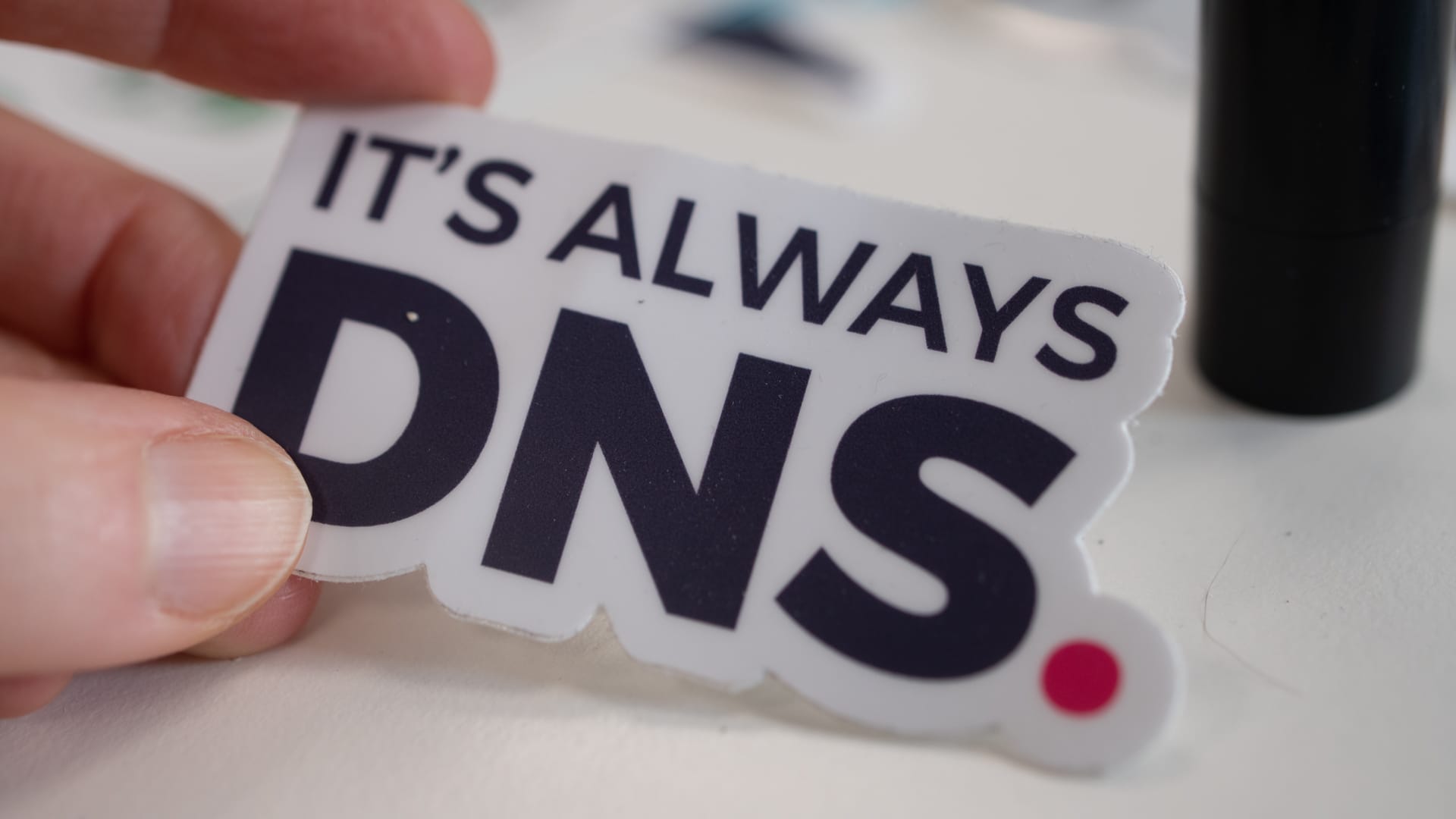 En hand som håller ett klistermärke som det står "it's always DNS" på.