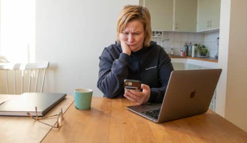 Kvinna sitter vid köksbord med dator framför sig, i handen håler hon mobilen, ser bekymrad ut.