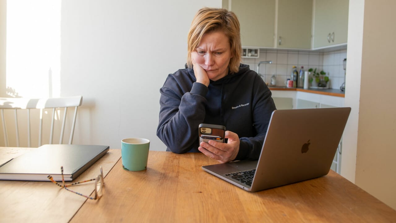 Kvinna sitter vid köksbord med dator framför sig, i handen håler hon mobilen, ser bekymrad ut.