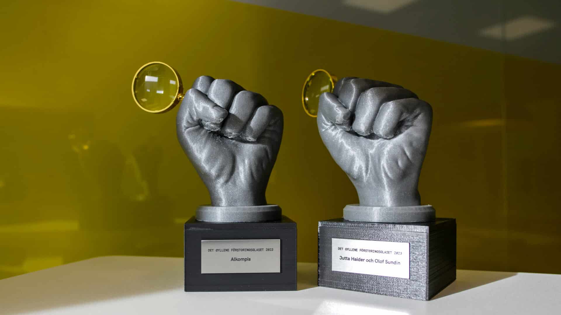 Två statyetter föreställandes gråa händer som håller gyllene förstoringsglas.