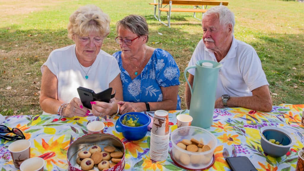 Tre äldre personer sitter vid ett bord med fika, tillsammans tittar de på en mobil.