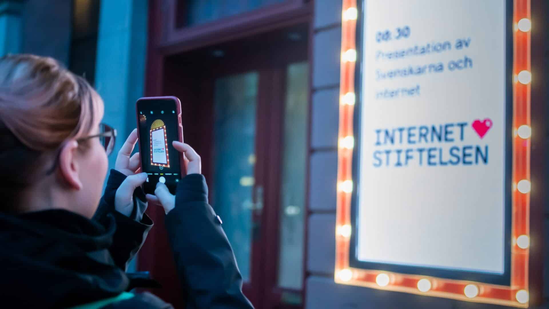 En kvinna står med en mobil och ta en bild på en reklamtavla, tavlan visar att rapporten Svenskarna och internet ska släppas.