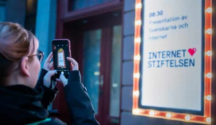 En kvinna står med en mobil och ta en bild på en reklamtavla, tavlan visar att rapporten Svenskarna och internet ska släppas.