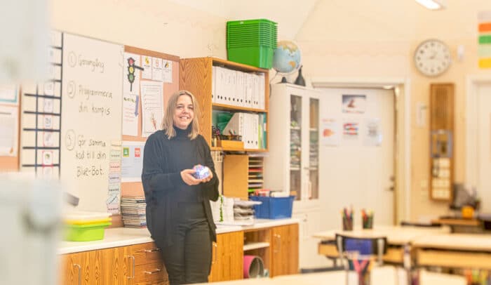 Lärare står i ett klassrum, i handen håller hon en robot som programmeras.