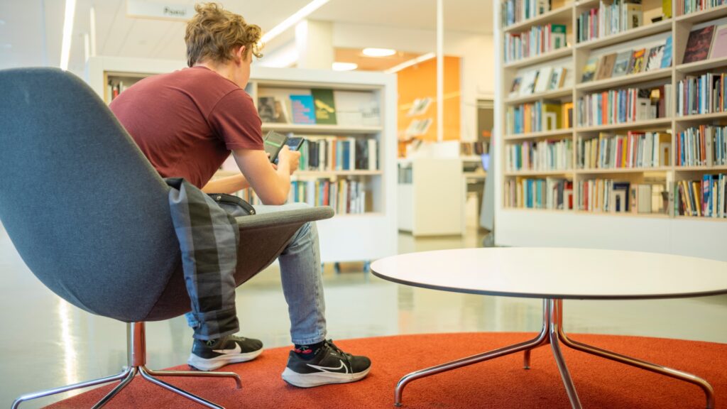 Hur går det med läsningen i Sverige?Ung kille sitter i biblioteket och använder sin mobiltelefon.