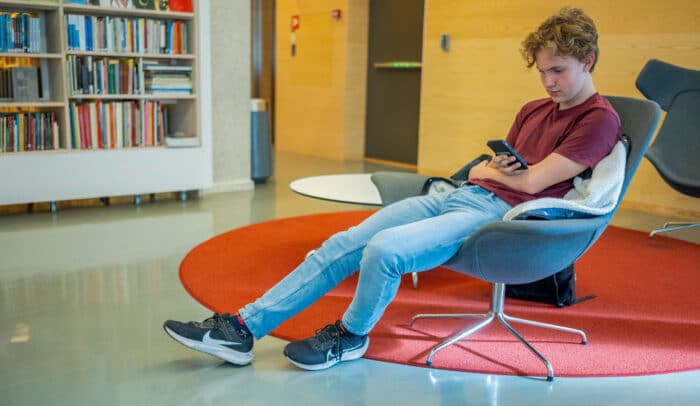 En ung man sitter på ett bibliotek och scrollar på sin mobil.