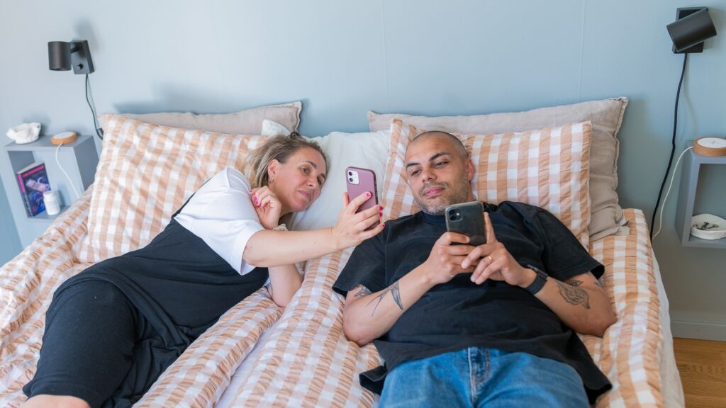 En man och en kvinna ligger i en säng med varsin mobil, kvinnan visar något för mannen.
