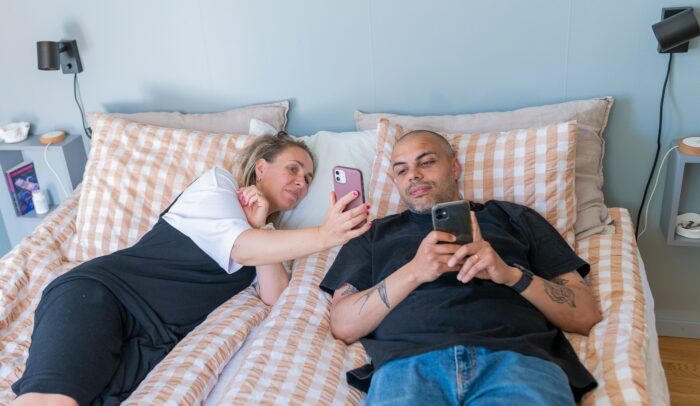 En man och en kvinna ligger i en säng med varsin mobil, kvinnan visar något för mannen.