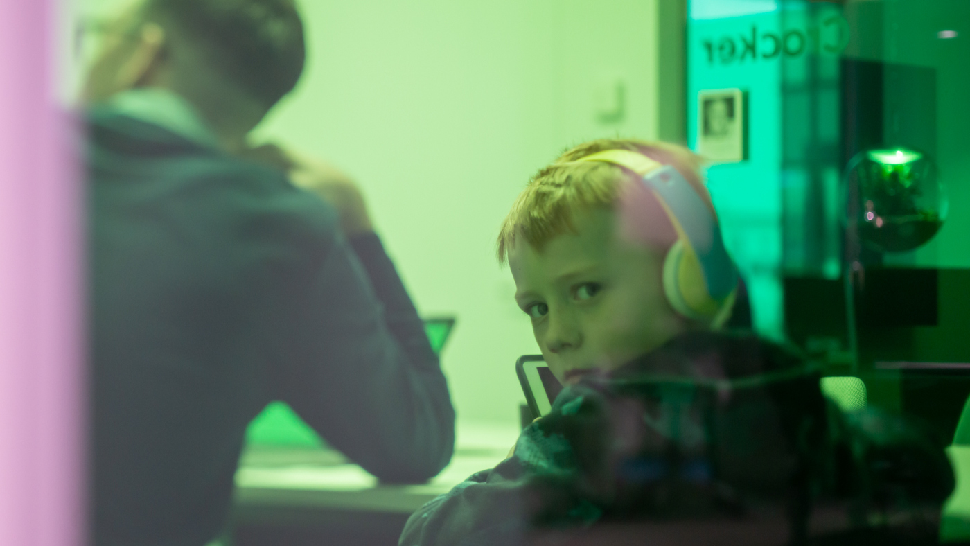 En pojke sitter framför en dator med hörlurar på sig, han tittar mot kameran.