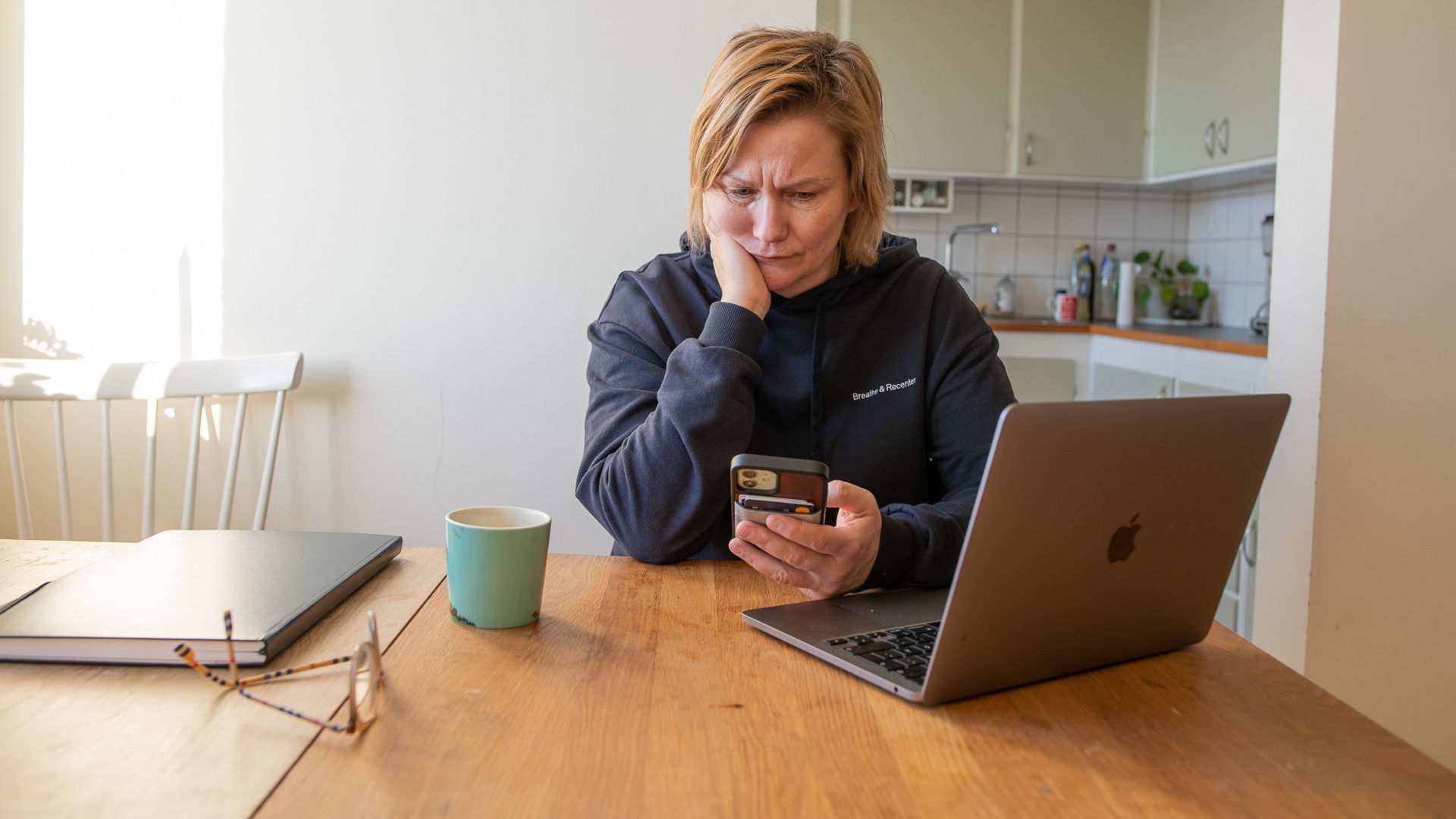 Ledsen person tittar på sin mobil, sittandes vid ett köksbord.