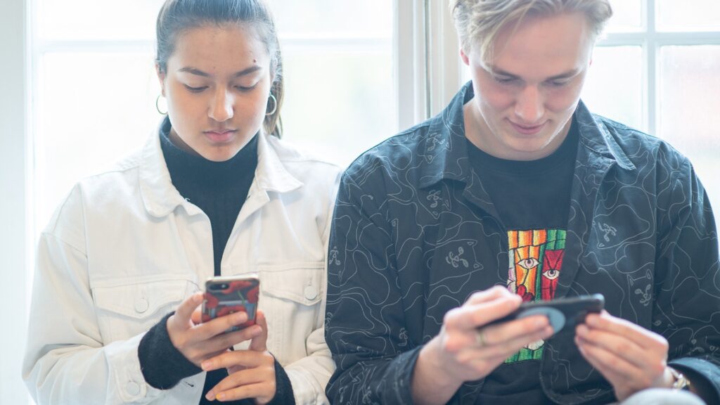 Två unga människor som tittar på sina mobiltelefoner. Artikeln handlar om trygga relationer på nätet för unga.