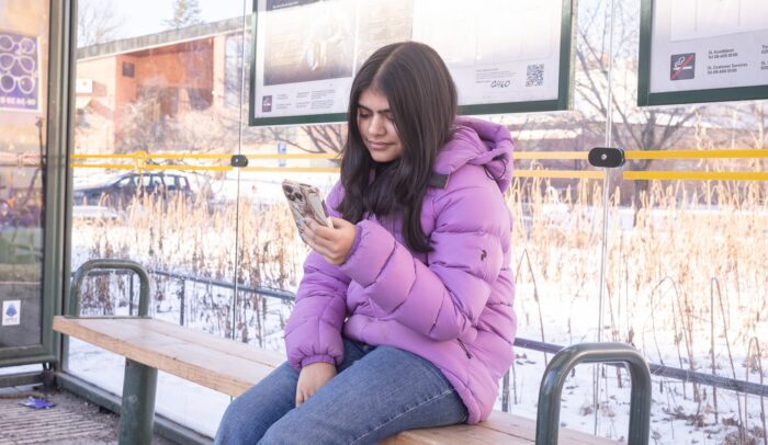 En ung tjej sitter på en busshållplats och scrollar på mobilen.