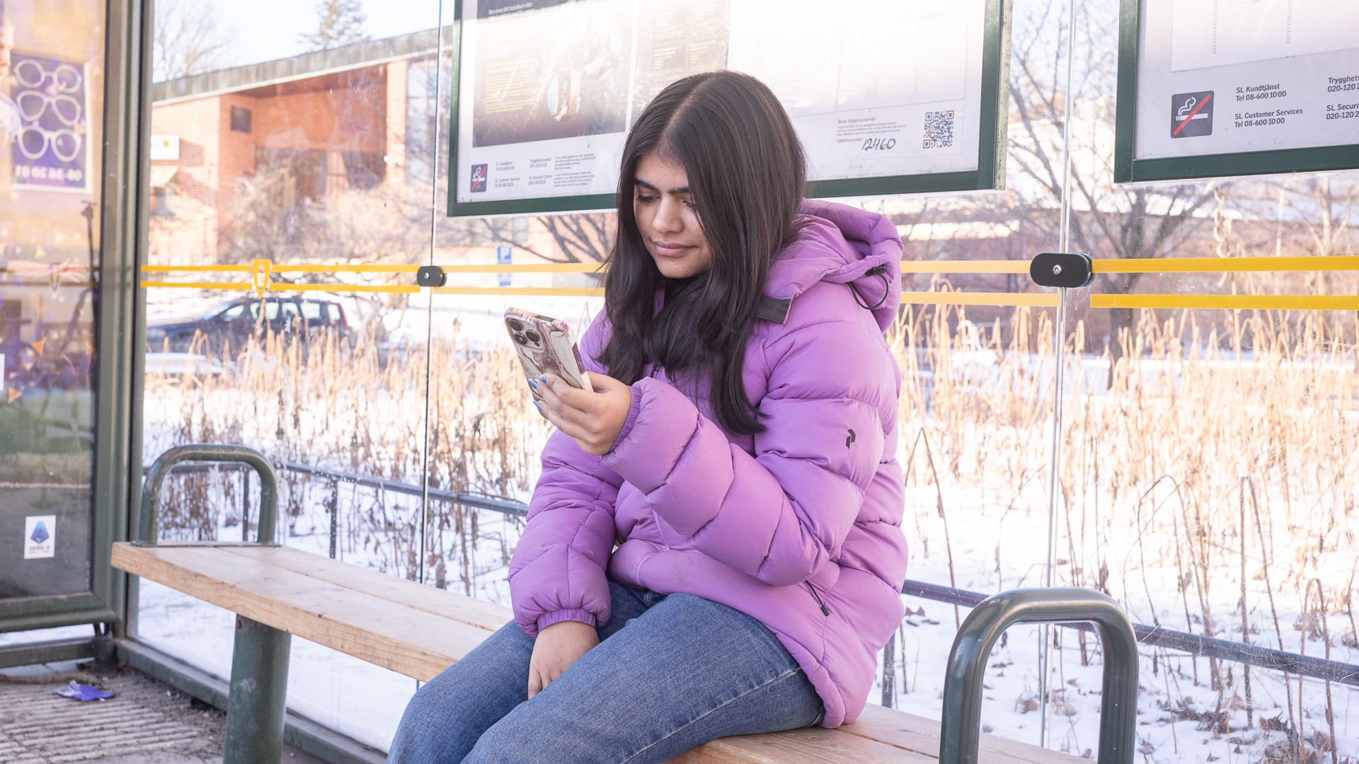 En ung tjej sitter på en busshållplats och scrollar på mobilen.