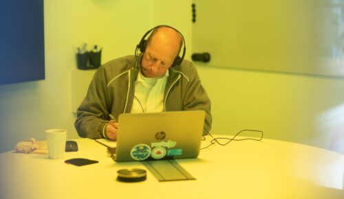 En man sitter i ett mötesrum med hörlurar på huvudet och en dator framför sig.