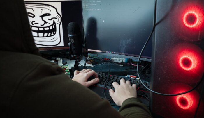 En person i svart hoodie sitter vid en dator, på skärmen syns ett otäckt ansikte.