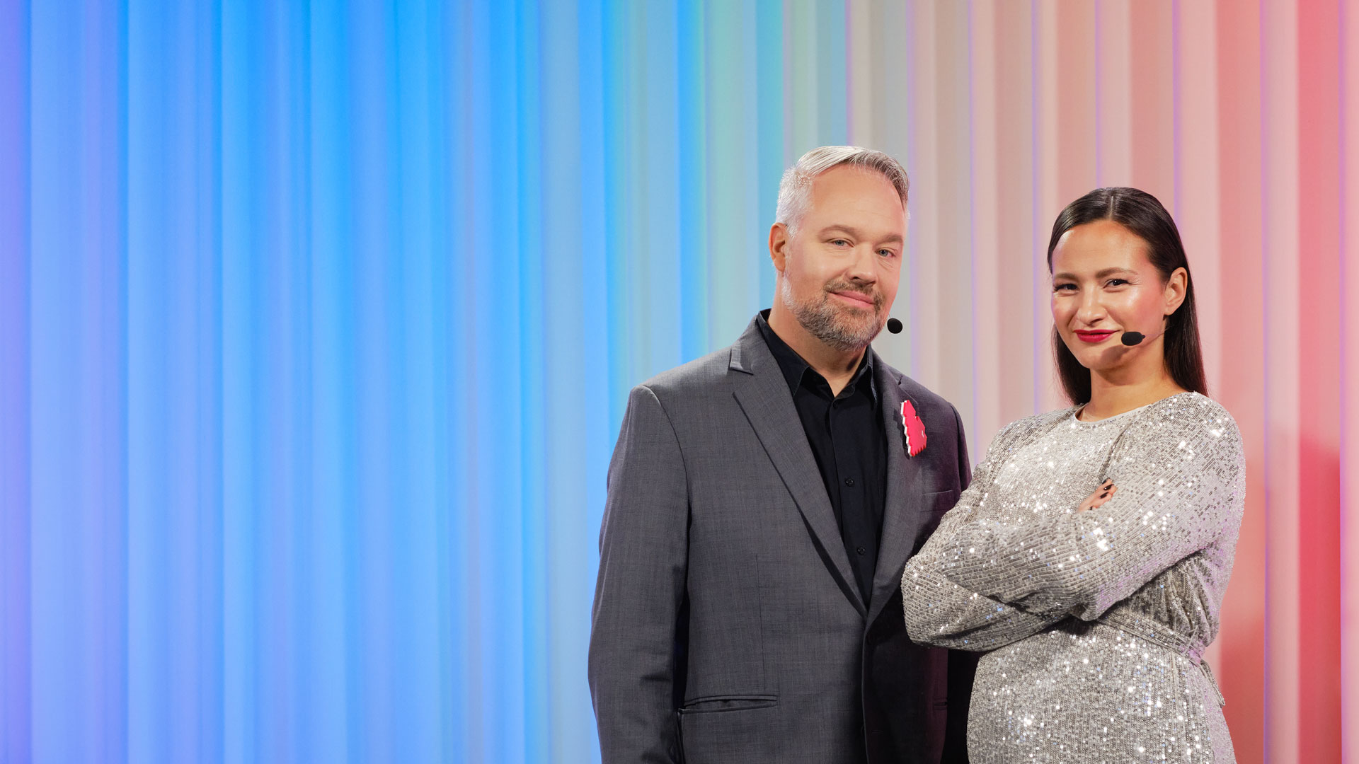 Två konferencierer, Måns Jonasson och Aisha Mossberg, står med huvudmikrofoner framför en färgad skärm.