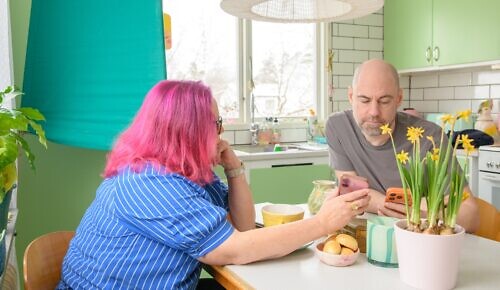 En man och en kvinna sitter vid köksbordet och ser på en mobiltelefon, framför en valskärm.