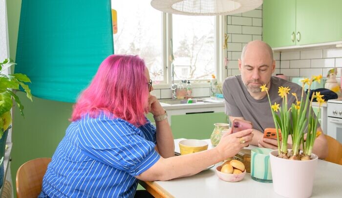 En man och en kvinna sitter vid köksbordet och ser på en mobiltelefon, framför en valskärm.