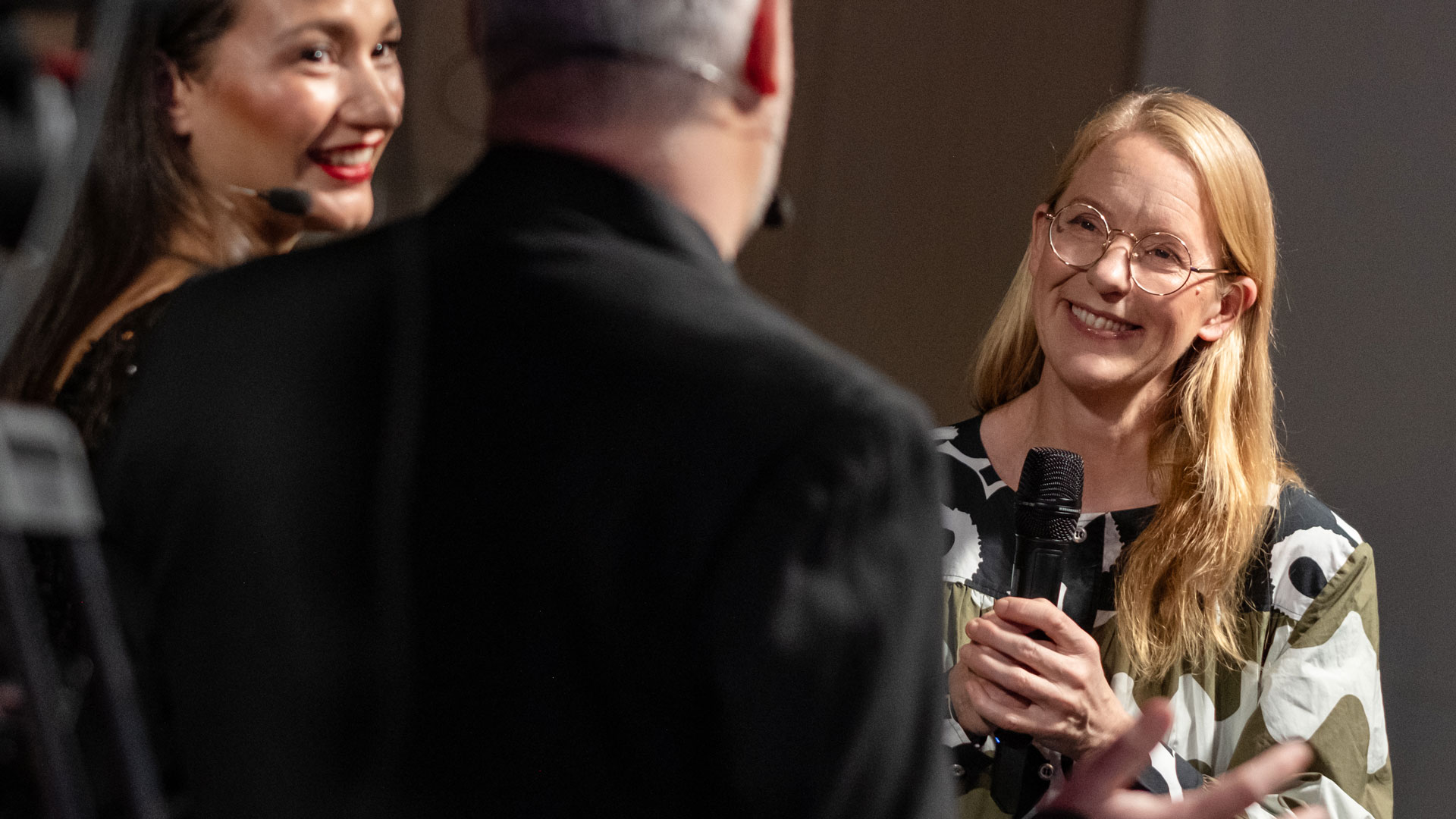 Två kvinnor med mikrofoner tittar på en tredje person som har ryggen mot kameran.