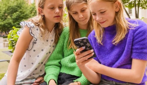 Tre flickor sitter på en bänk och tittar på en mobiltelefon.