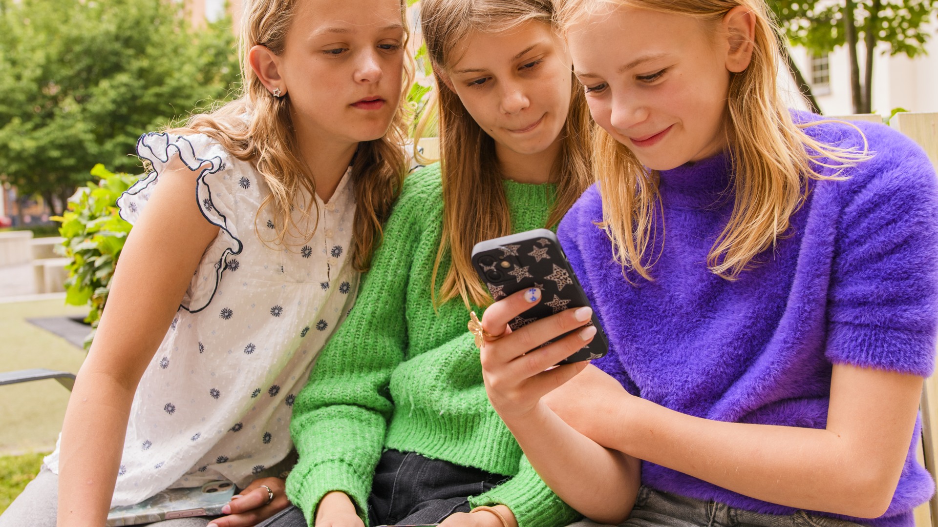 Tre flickor sitter på en bänk och tittar på en mobiltelefon.