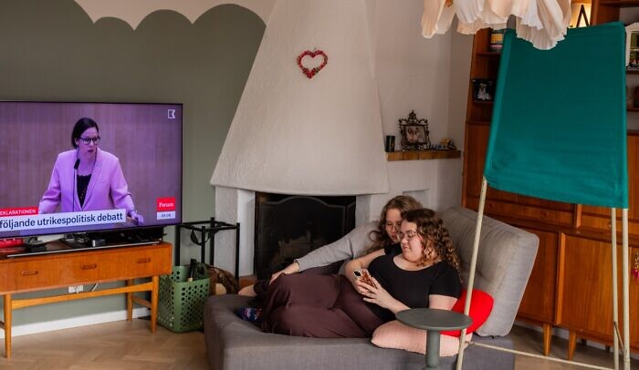 Två kvinnor följer en tv-debatt och kollar på sin mobiltelefon. Bakom dom står ett valbås.