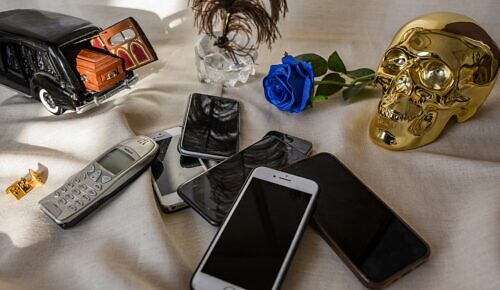 Många mobiltelefoner av olika ålder ligger på ett bord bredvid en leksaksbil med en likkista i.