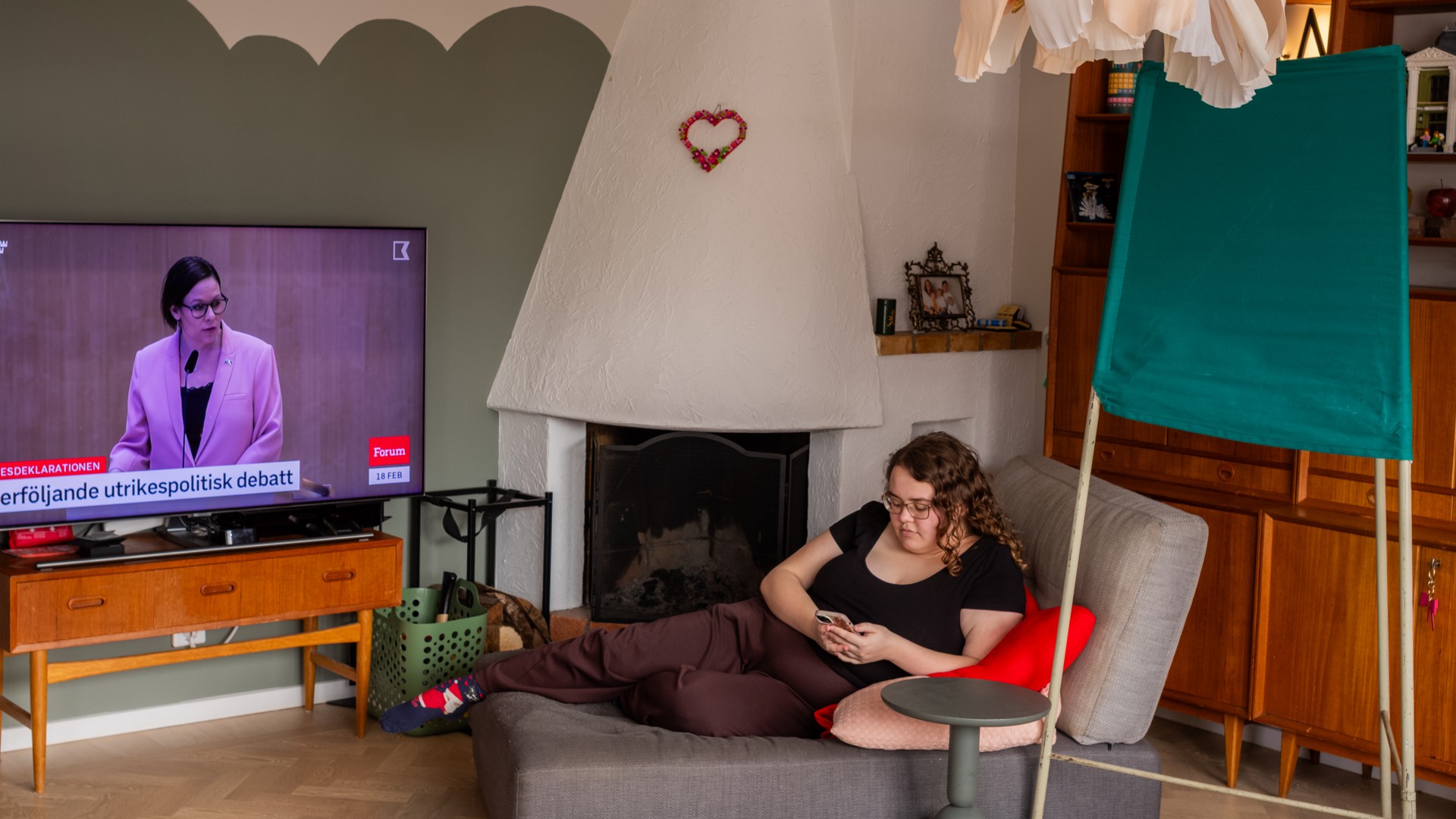 Kvinna ligger i soffa och kollar på sin mobil. Bredvid soffan står en tv och en vallokalsskärm.